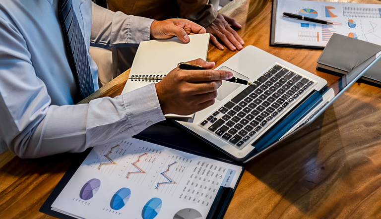 Administradores em reunião diante de laptop, analisando planilhas e cadernos.