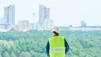 Profissional com capacete e colete de proteção observa floresta com prédios ao fundo.