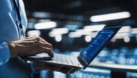 Homem com camisa social e crachá trabalha em notebook.