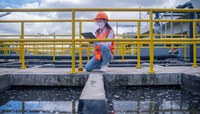 Mulher profissional com prancheta e equipamentos de proteção avalia amostra de líquido/água de tanque industrial.