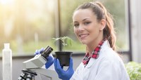 Mulher com jaleco branco e luvas sorri para a câmera enquanto segura vaso de planta e microscópio.