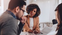 Duas mulheres e um homem vestidos com roupa social analisam documentos em reunião de negócios.