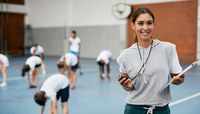 Professora de educação física com cronômetro e prancheta e roupas de esporte. Ao fundo alunos se alongando em quadra.