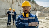 Técnico com equipamentos de proteção individual em mina com trabalhadores e trator ao fundo.